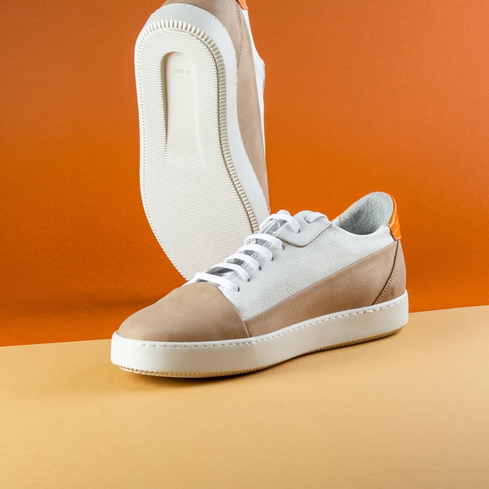 Prodcut photo of a pair of sneakers placed on a orange background. The sneakers have a white sole, a brown-white shoe and a orange heel.